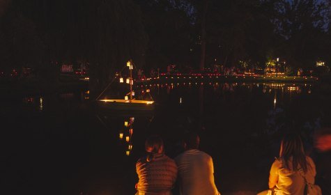 Im Vordergrund sind ein Paar und eine einzelne Person zu sehen, die bei der Nacht der 10.000 Kerzen auf den kleinen Kurparksee schauen, auf dem See schwimmt ein Boot mit Kerzen, © Stadt Bad Salzuflen | Nadja Jacke