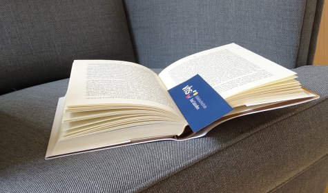 Auf einem Sofa liegt ein aufgeschlagenes Buch, in ihm steckt ein Lesezeichen mit Logo der VHS Bad Salzuflen, © Stadt Bad Salzuflen