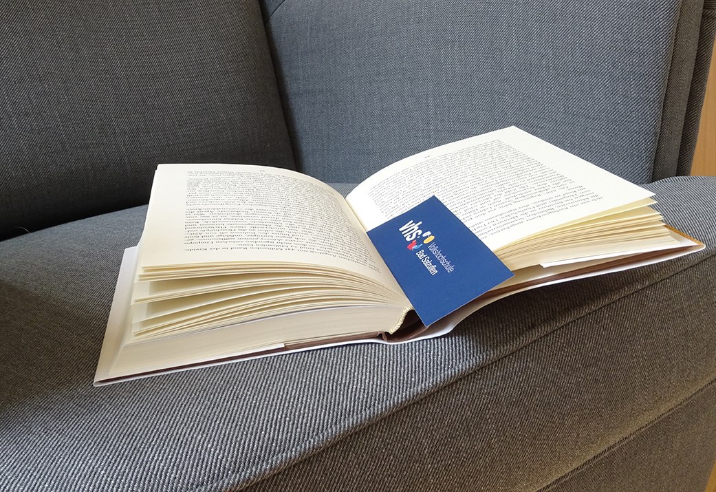 Auf einem Sofa liegt ein aufgeschlagenes Buch, in ihm steckt ein Lesezeichen mit Logo der VHS Bad Salzuflen, © Stadt Bad Salzuflen