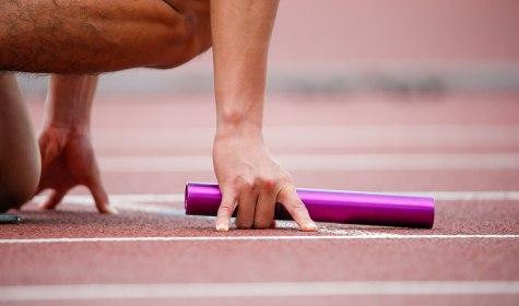 Läufer kniet in Startposition auf der Tartanbahn und hält einen Staffelstab in der Hand., © stock.adobe.com -  D maborosi