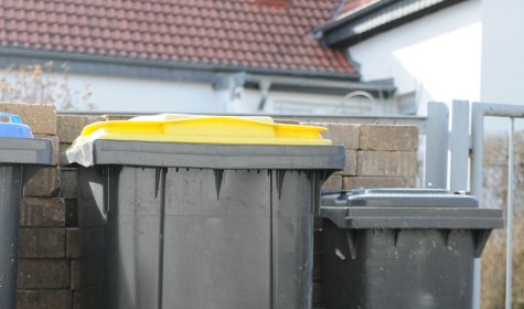 Auf dem Bild zu sehen ist eine Mauer, vor der verschiedene Mülltonnen stehen. Im Mittelpunkt des Bildes steht eine schwarze Mülltonne mit gelbem Deckel. Es handelt sich um die Gelbe Tonne für Leichtverpackungen., © fotoak80 - stock.adobe.com